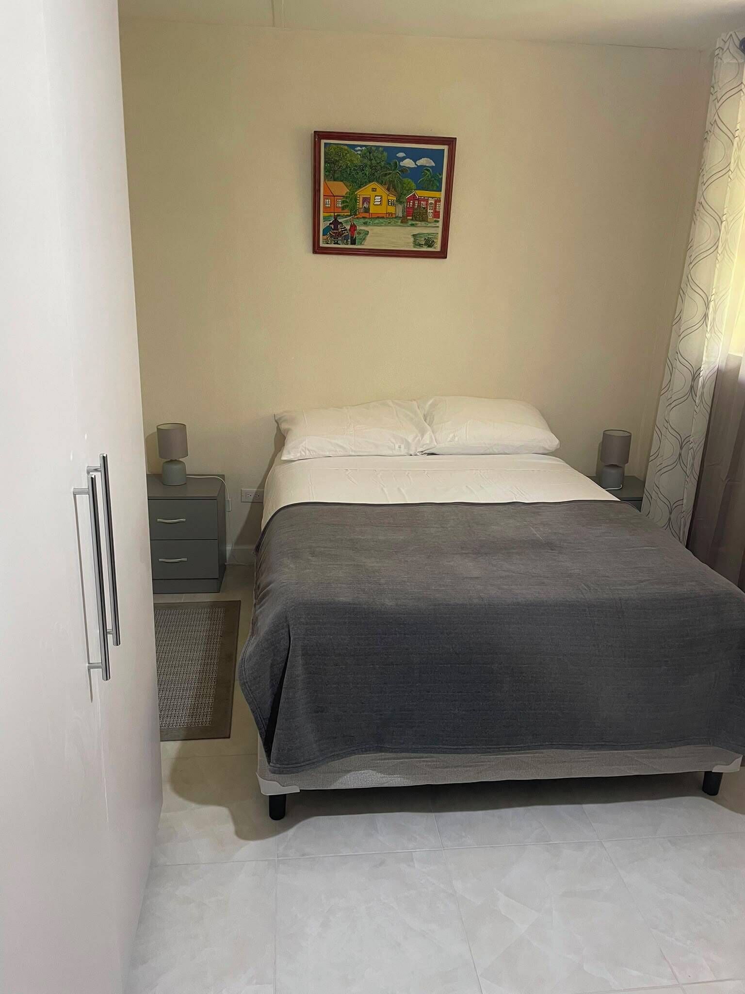 A bedroom with a bed, a nightstand, a drawer unit, a painting on the wall, and a curtain.