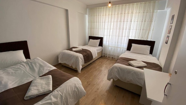 A hotel room with three single beds, a wooden floor, a window with sheer curtains, and a small bedside table with a lamp.