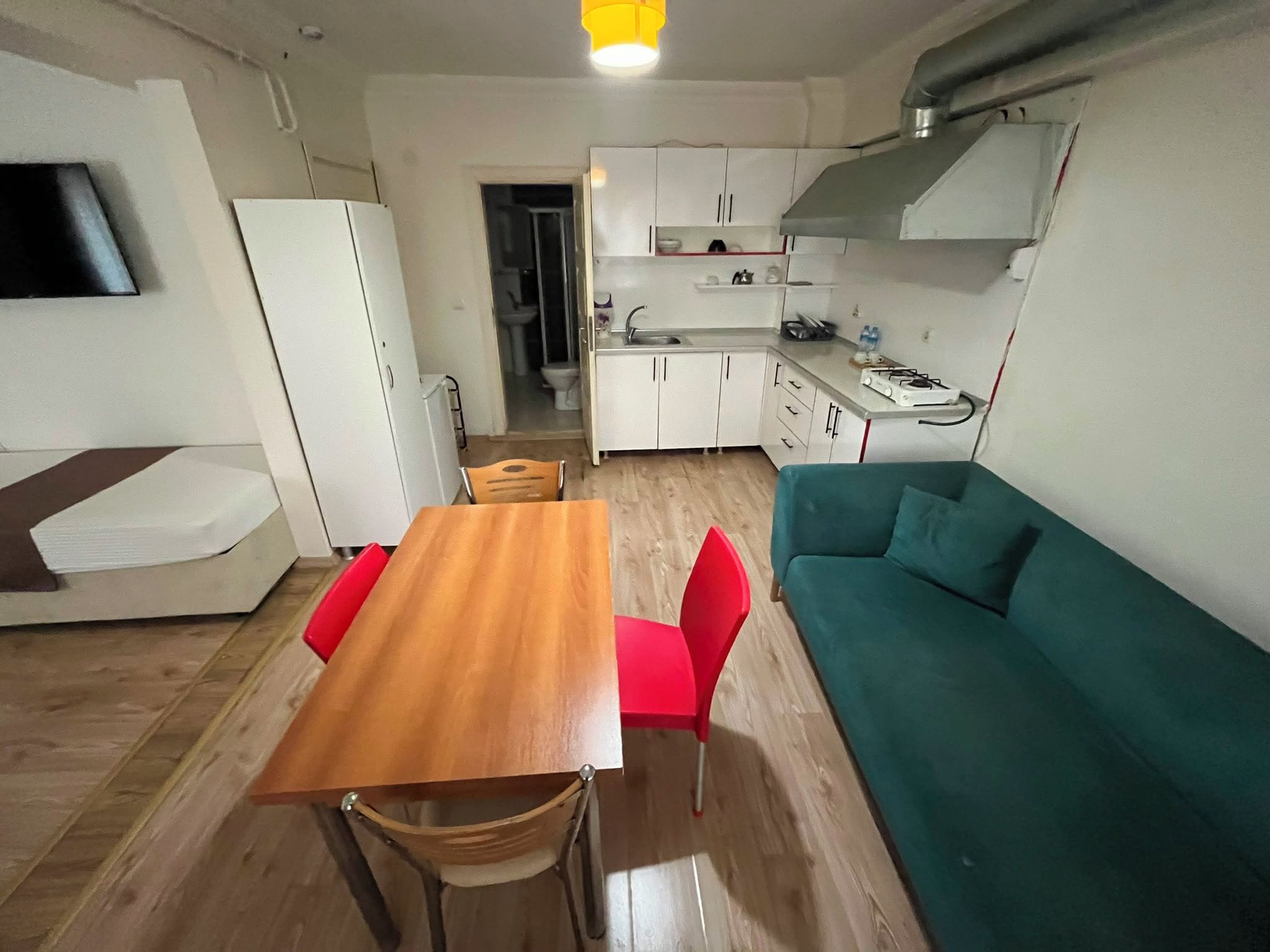 Um espaço de estar compacto com cozinha compacta, área de jantar com mesa de madeira e cadeiras vermelhas, e sofá azul-esverdeado.