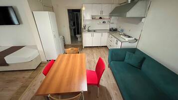 Um espaço de estar compacto com cozinha compacta, área de jantar com mesa de madeira e cadeiras vermelhas, e sofá azul-esverdeado.