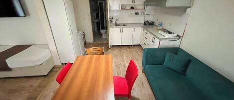 A compact living space with a kitchenette, a dining area with a wooden table and red chairs, and a teal sofa.