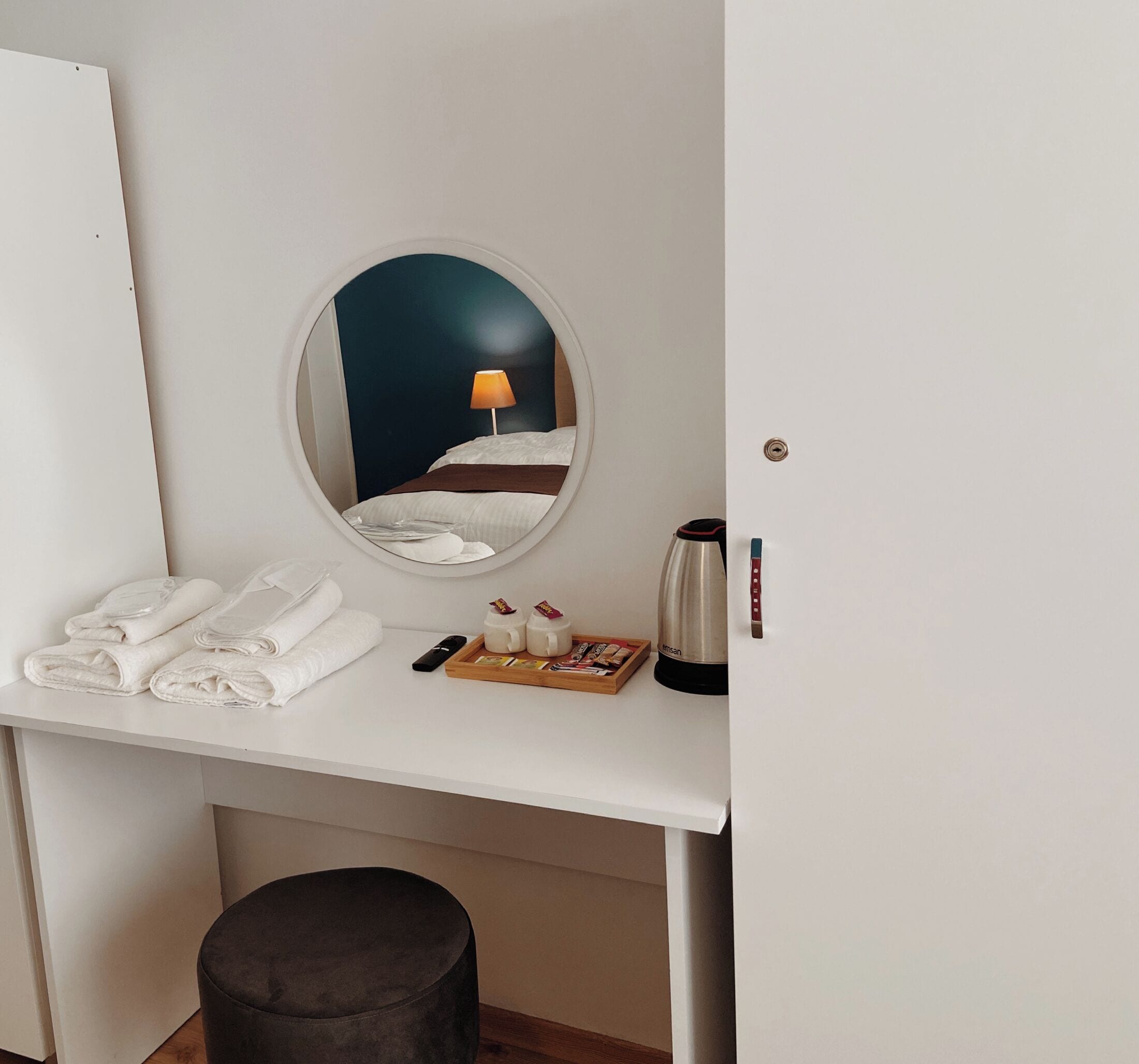 Quarto de hotel com um espelho redondo, uma cama com roupa de cama branca, uma mesa de cabeceira com abajur, uma chaleira e um suporte com lanches.
