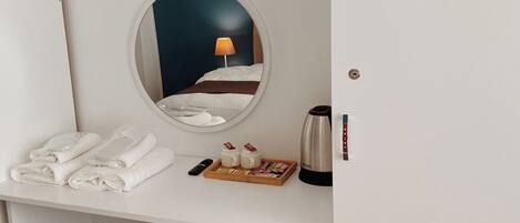 Une chambre d’hôtel avec un miroir rond, un lit avec du linge de lit blanc, une table de chevet avec une lampe, une bouilloire et un plateau avec des en-cas.