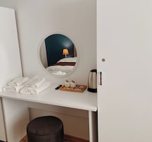 A hotel room with a round mirror, a bed with white linens, a bedside table with a lamp, a kettle, and a tray with snacks.
