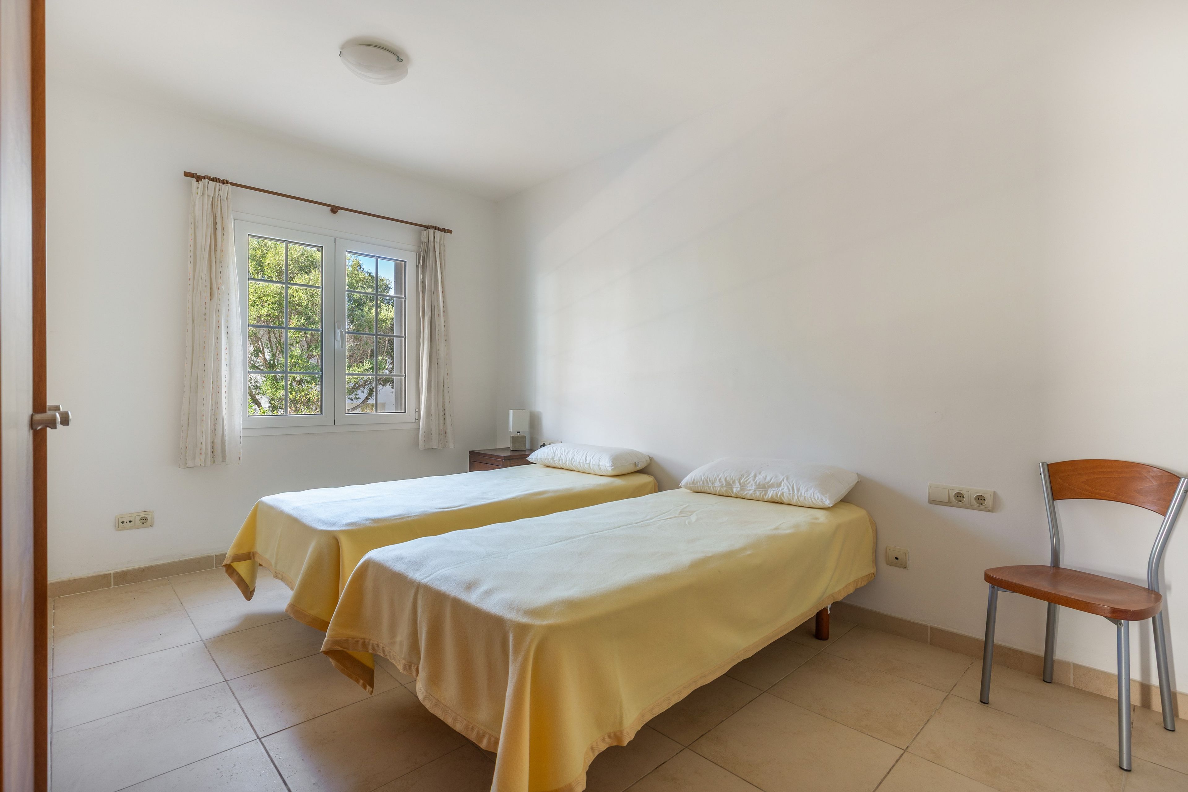 Ein Zimmer mit zwei Einzelbetten, einem Sessel, einem Fenster mit Vorhängen und einem Nachttisch mit einer Lampe.