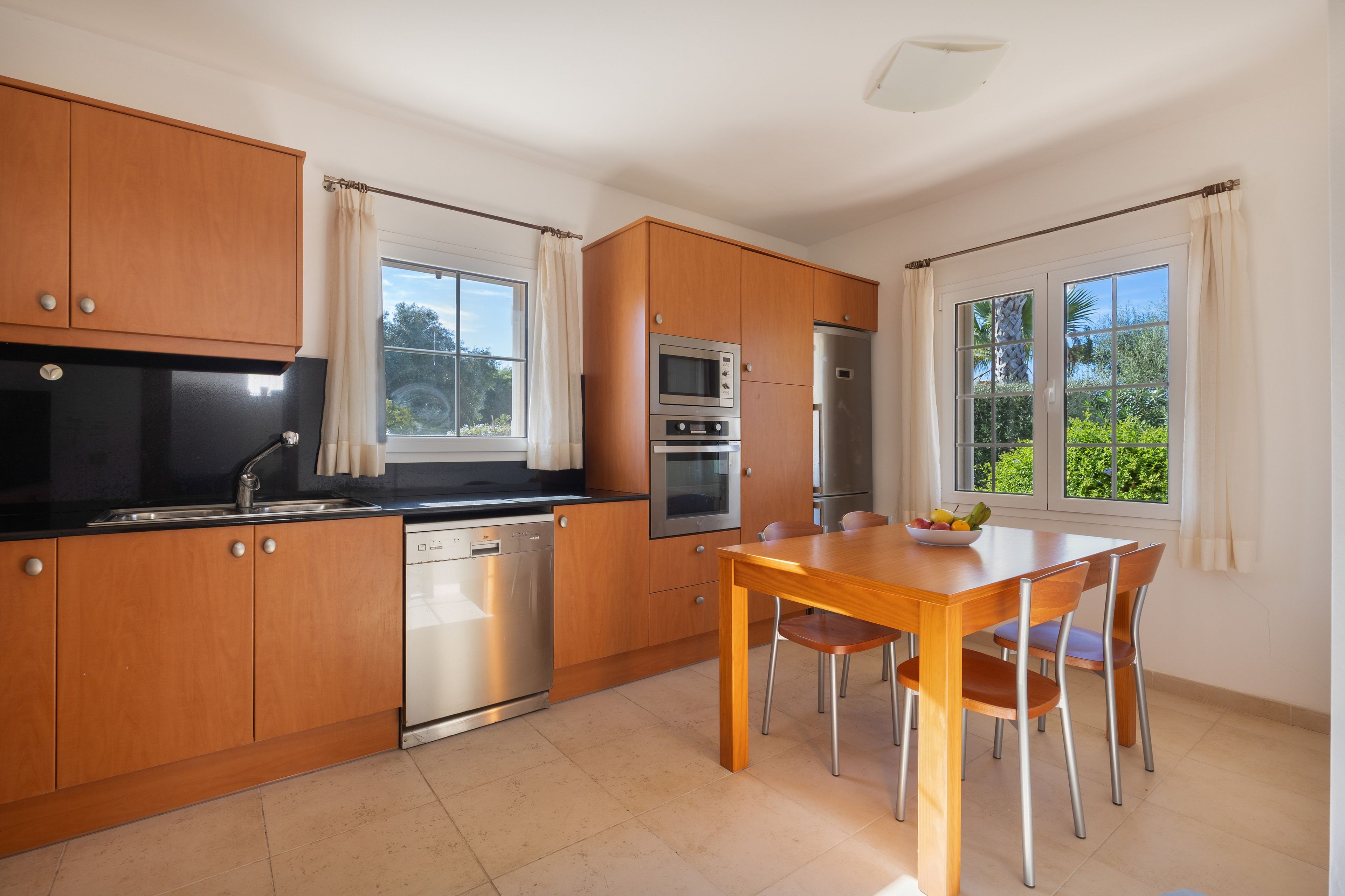 Cocina moderna con armarios de madera, lavavajillas, estufa, horno y refrigerador. Hay una mesa de comedor con sillas y un cuenco de fruta.