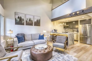 Una sala moderna con un sofá gris, un reposapiés marrón y una mesa de centro. Se observa una zona de cocina con un refrigerador de acero inoxidable y una barra.