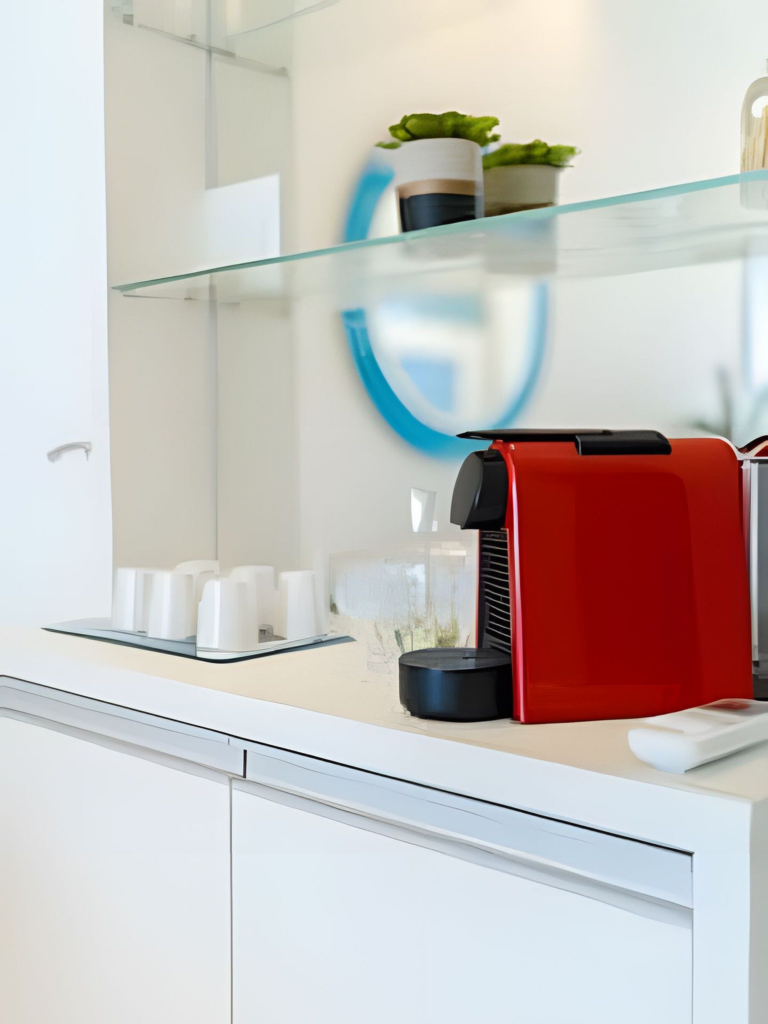 Une salle de bain moderne avec une étagère en verre où sont rangés les articles de toilette, une machine à café rouge et un lavabo blanc.