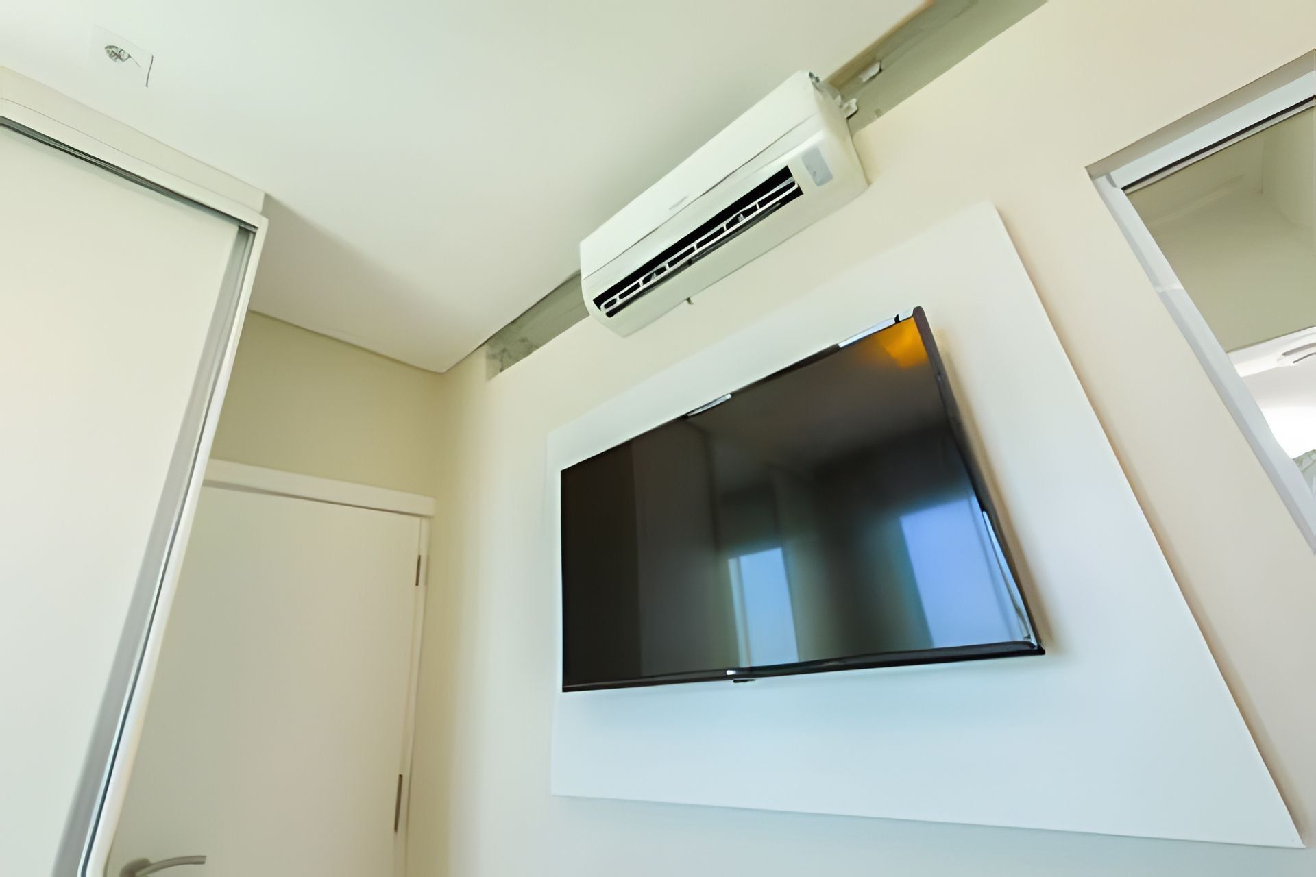 Una habitación con un televisor de pantalla plana montado, una puerta blanca y un equipo de aire acondicionado en el techo.