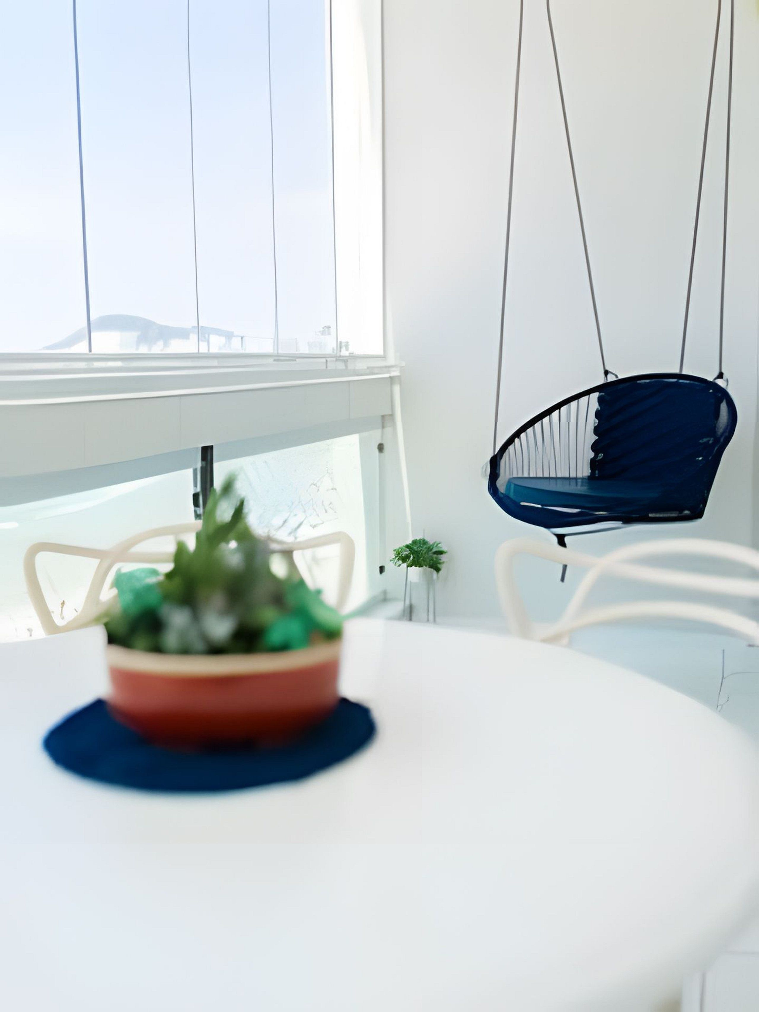 Uma mesa branca com uma tigela vermelha cheia de plantas verdes, uma cadeira azul pendurada e vista para edifícios através de grandes janelas.