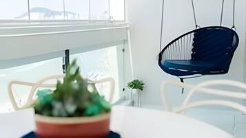 Uma mesa branca com uma tigela vermelha cheia de plantas verdes, uma cadeira azul pendurada e vista para edifícios através de grandes janelas.