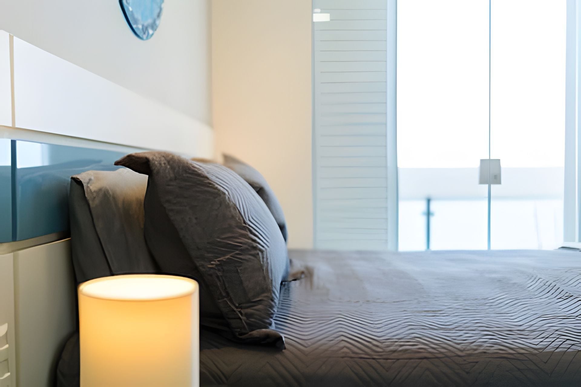 Quarto moderno com cama, almofadas, abajur e vista para o mar através de amplas janelas.