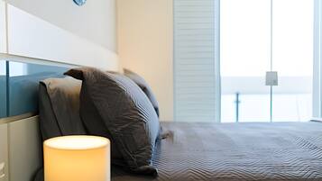 Quarto moderno com cama, almofadas, abajur e vista para o mar através de amplas janelas.