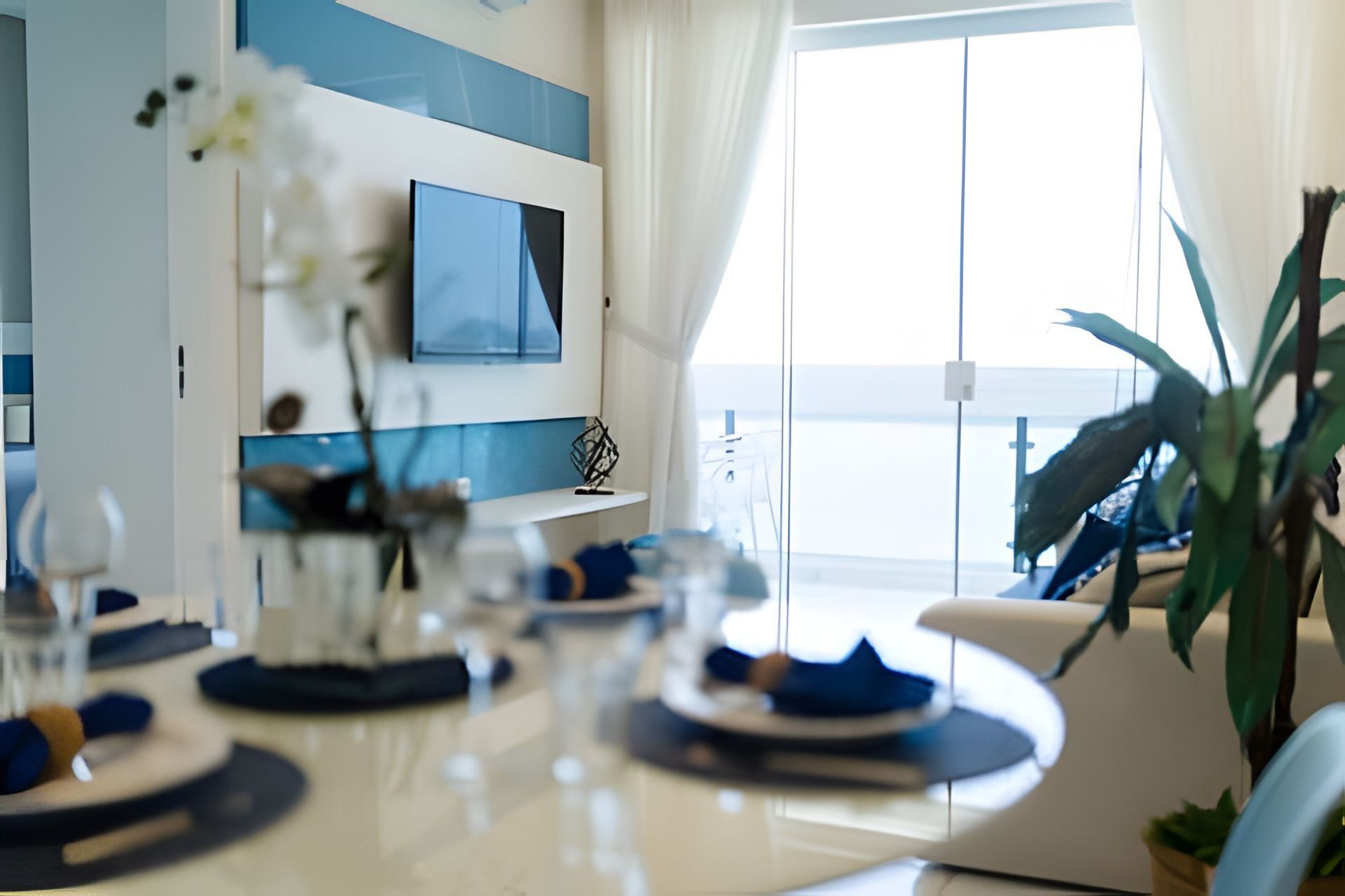 A modern dining area with a white table set for four, a flat-screen TV mounted on the wall, and a view of the sea through large windows.