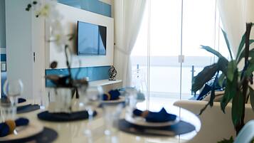 A modern dining area with a white table set for four, a flat-screen TV mounted on the wall, and a view of the sea through large windows.