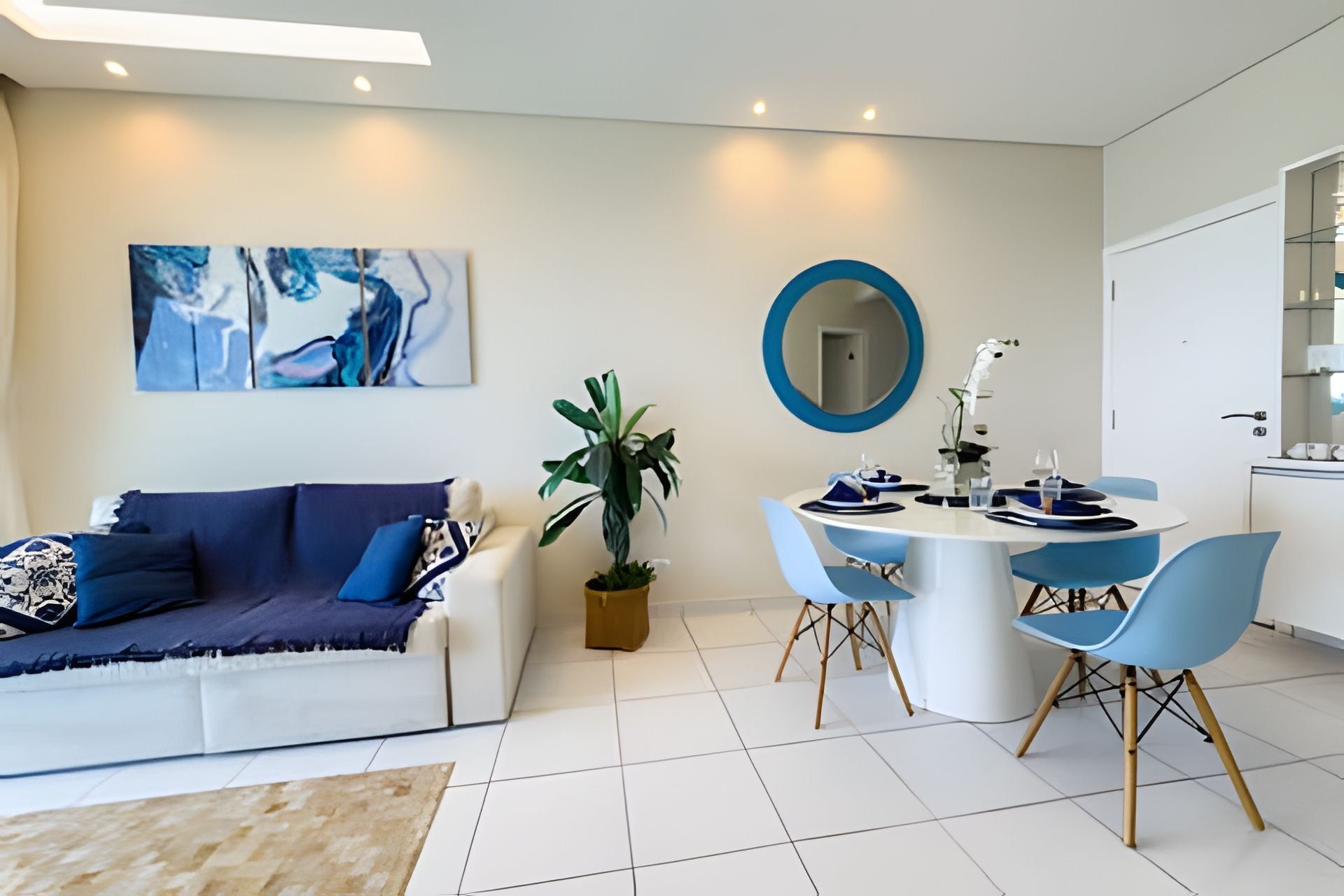 A modern living room with a white sofa, blue cushions, a coffee table, and a dining area with a round table and blue chairs.