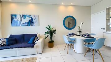 A modern living room with a white sofa, blue cushions, a coffee table, and a dining area with a round table and blue chairs.