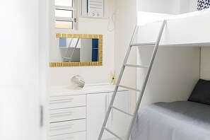 Una cama litera con escalera, un aparador blanco con cajones, un espejo y una ventana con persianas.