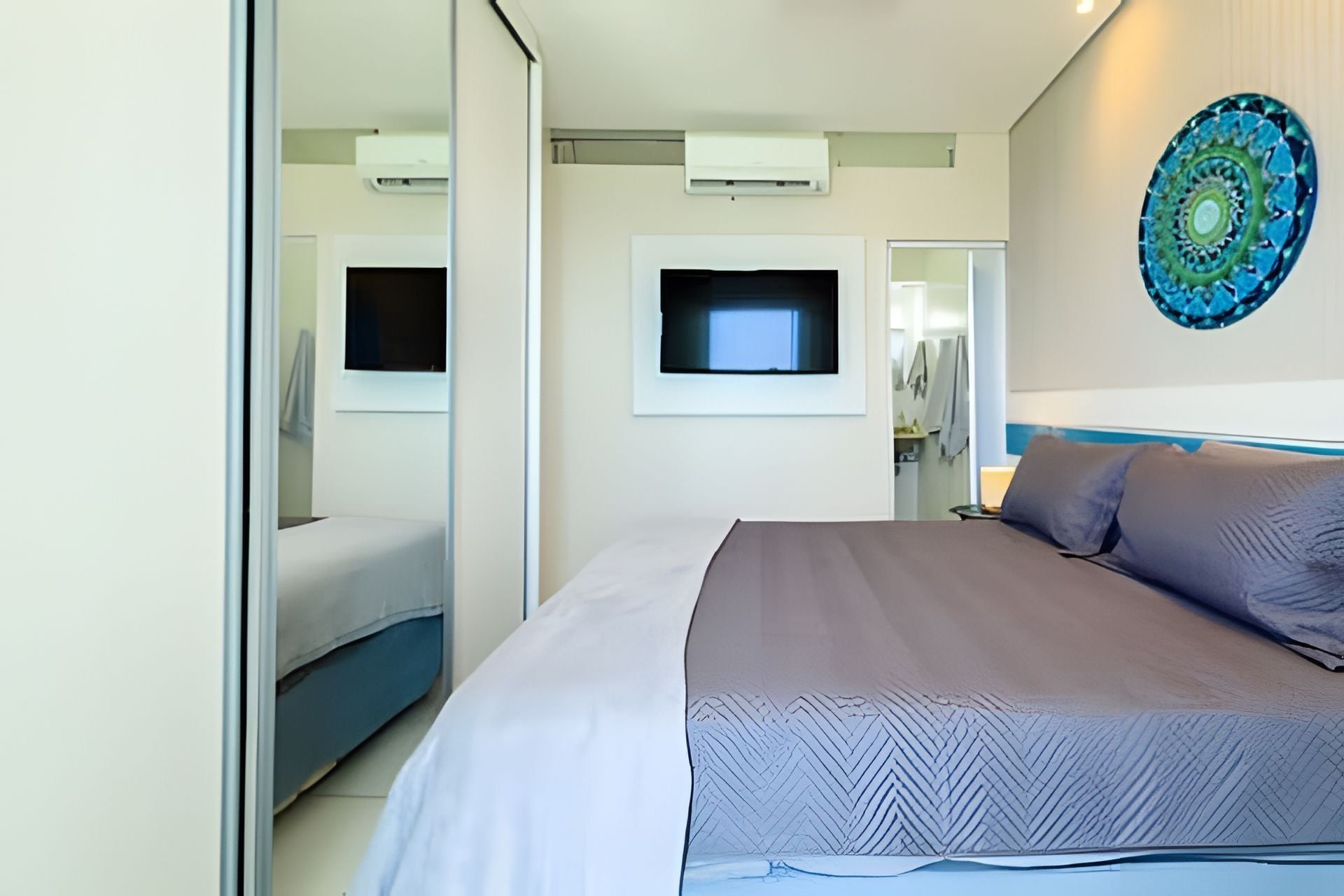 Quarto de hotel moderno com uma cama grande, uma televisão de tela plana na parede e um espelho decorativo.