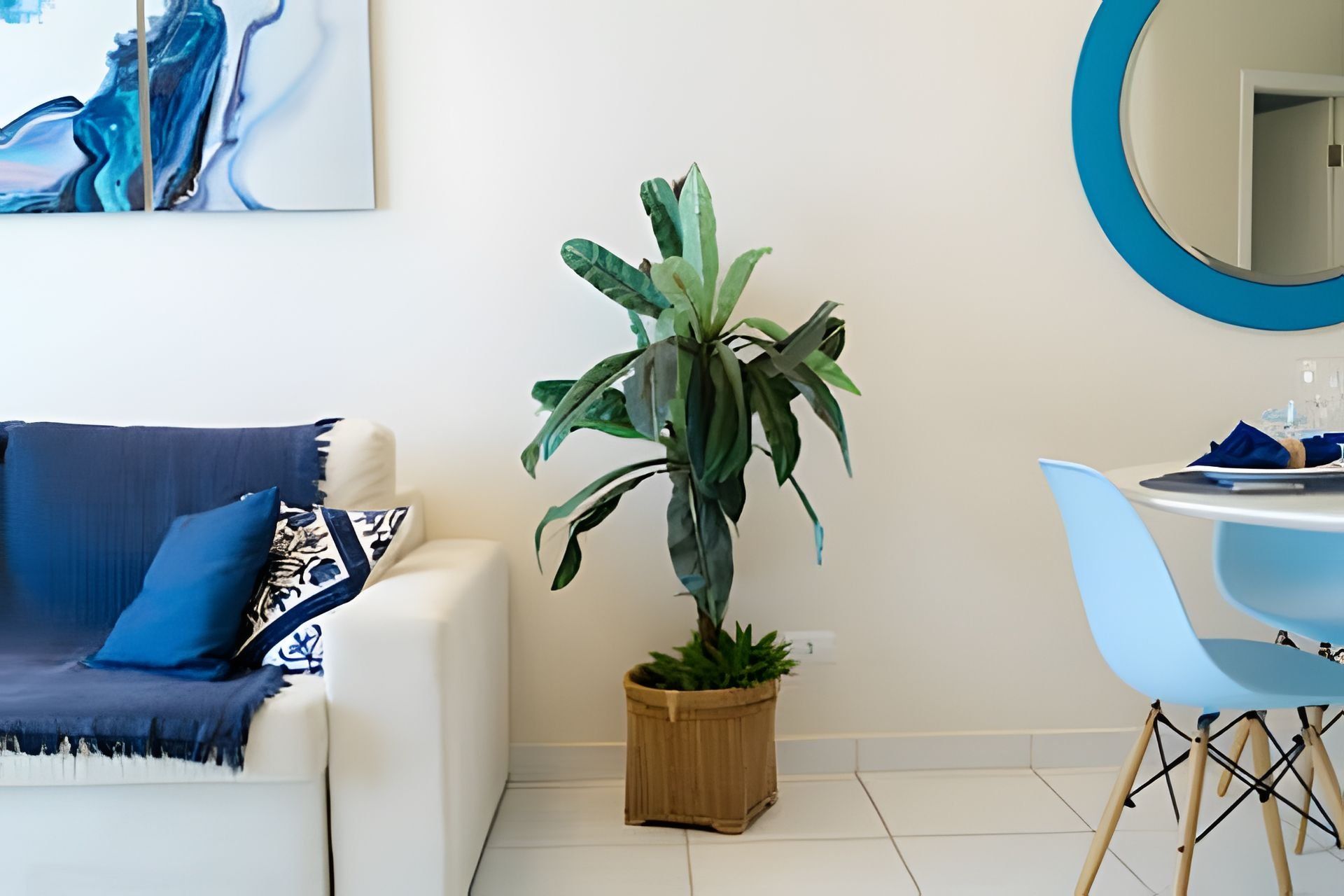 Uma sala de estar com um sofá branco, uma almofada azul, um vaso de planta de madeira, uma cadeira azul e um espelho redondo na parede.