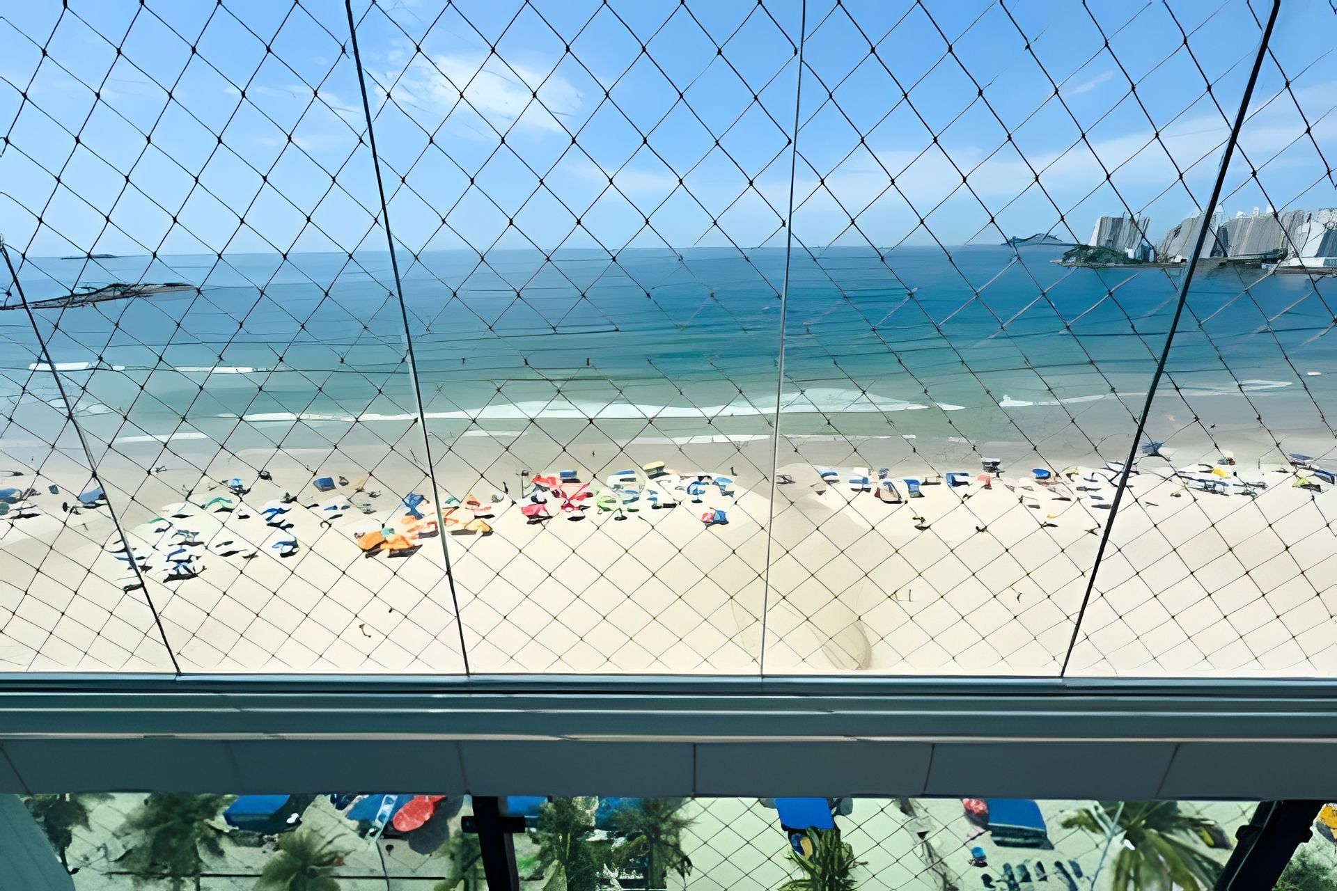 A beach view through a chain-link fence with a clear sky, sandy beach, and several umbrellas.