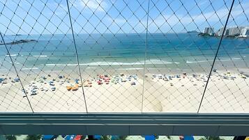 A beach view through a chain-link fence with a clear sky, sandy beach, and several umbrellas.