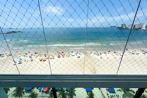 Vista de una playa a través de una cerca de malla metálica, con cielo despejado, arena y varios paraguas.