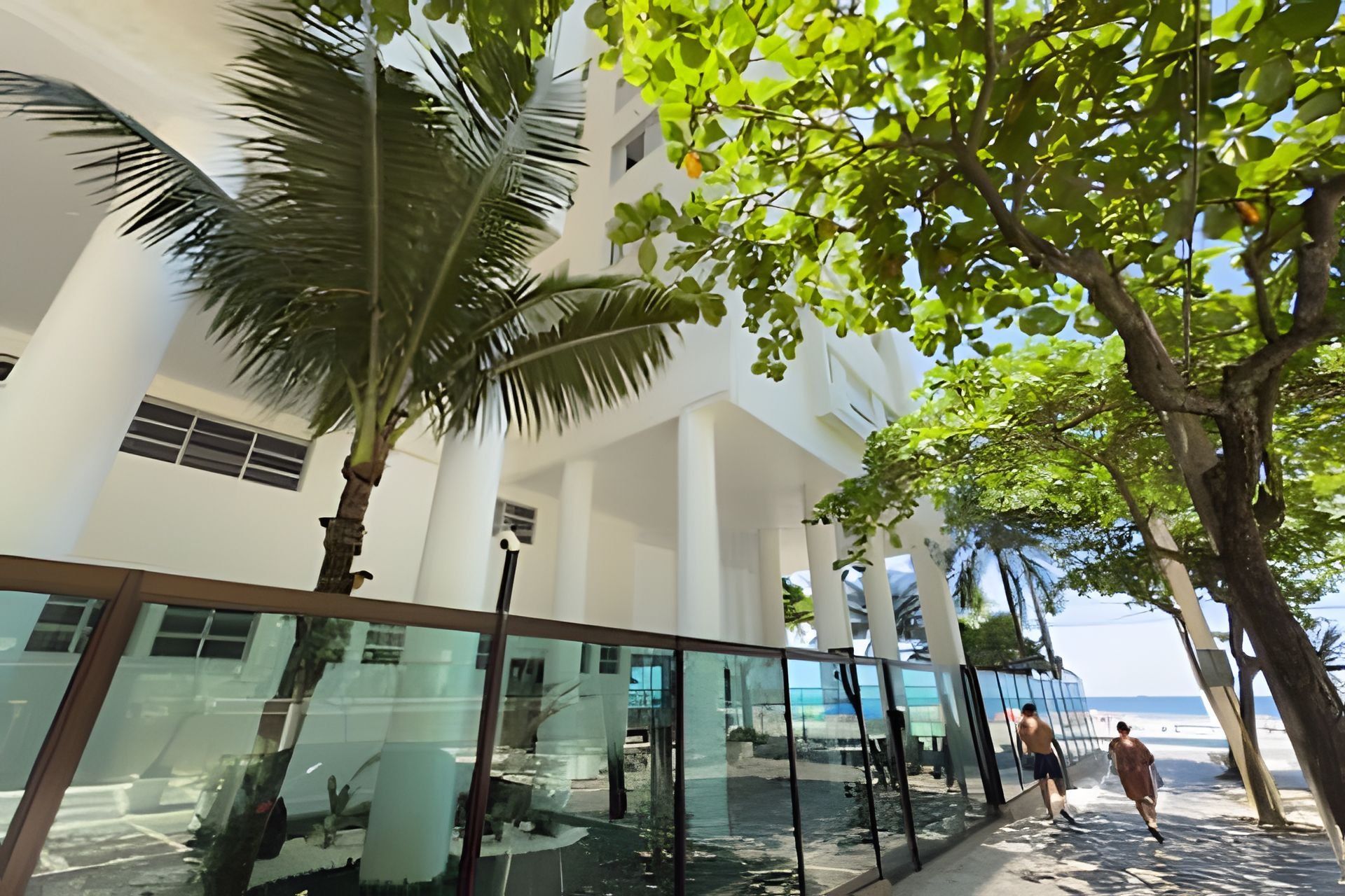 Un hotel moderno con un pasillo de vidrio, palmeras y vista a la playa.