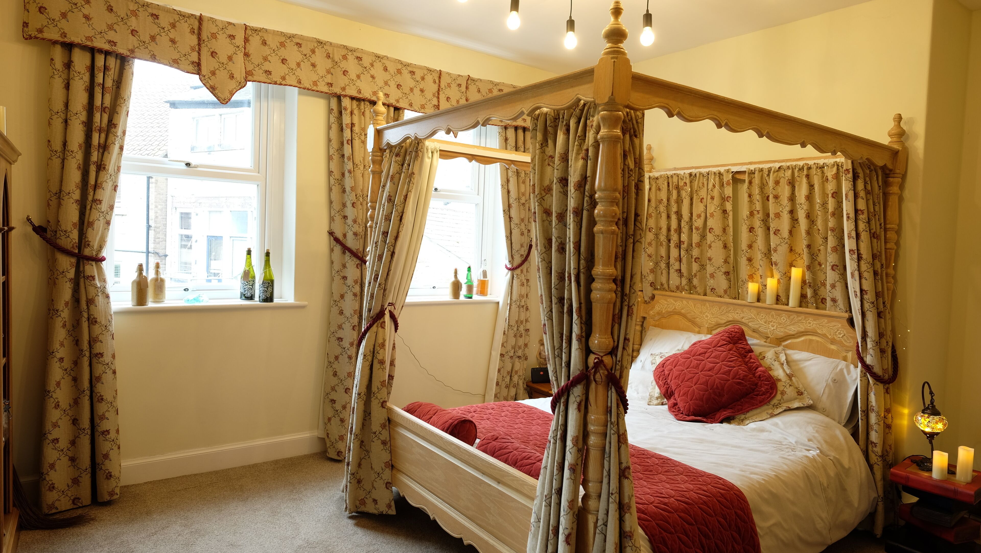 A four-poster canopy bed with a red quilt, a bedside table with candles, and a window with bottles.