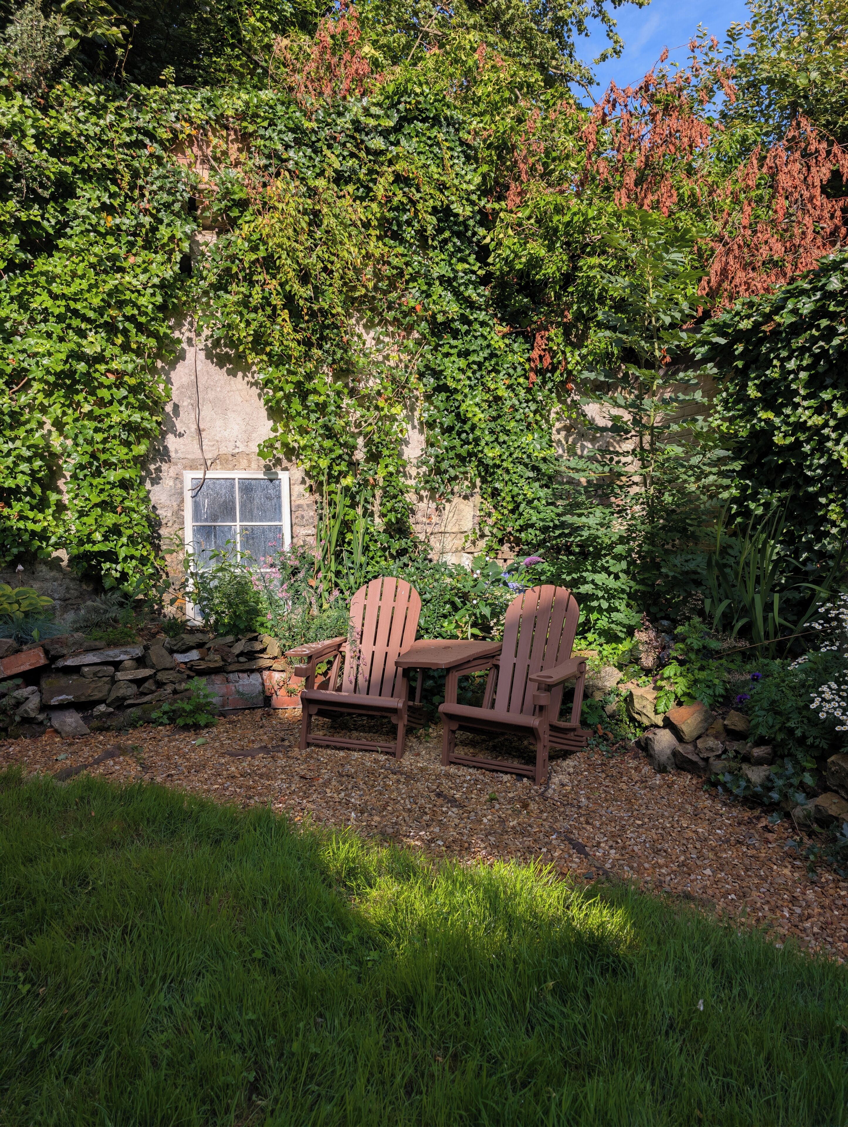 A garden with a stone wall, two Adirondack chairs, and a window.
