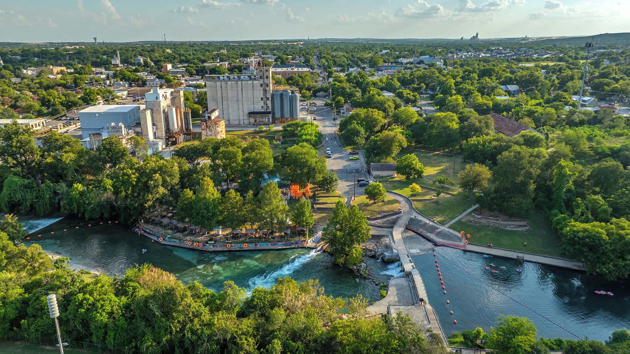 Ein Park am Flussufer mit einer Bootsanlegestelle, einer Brücke und einem großen Industriegebäude im Hintergrund.