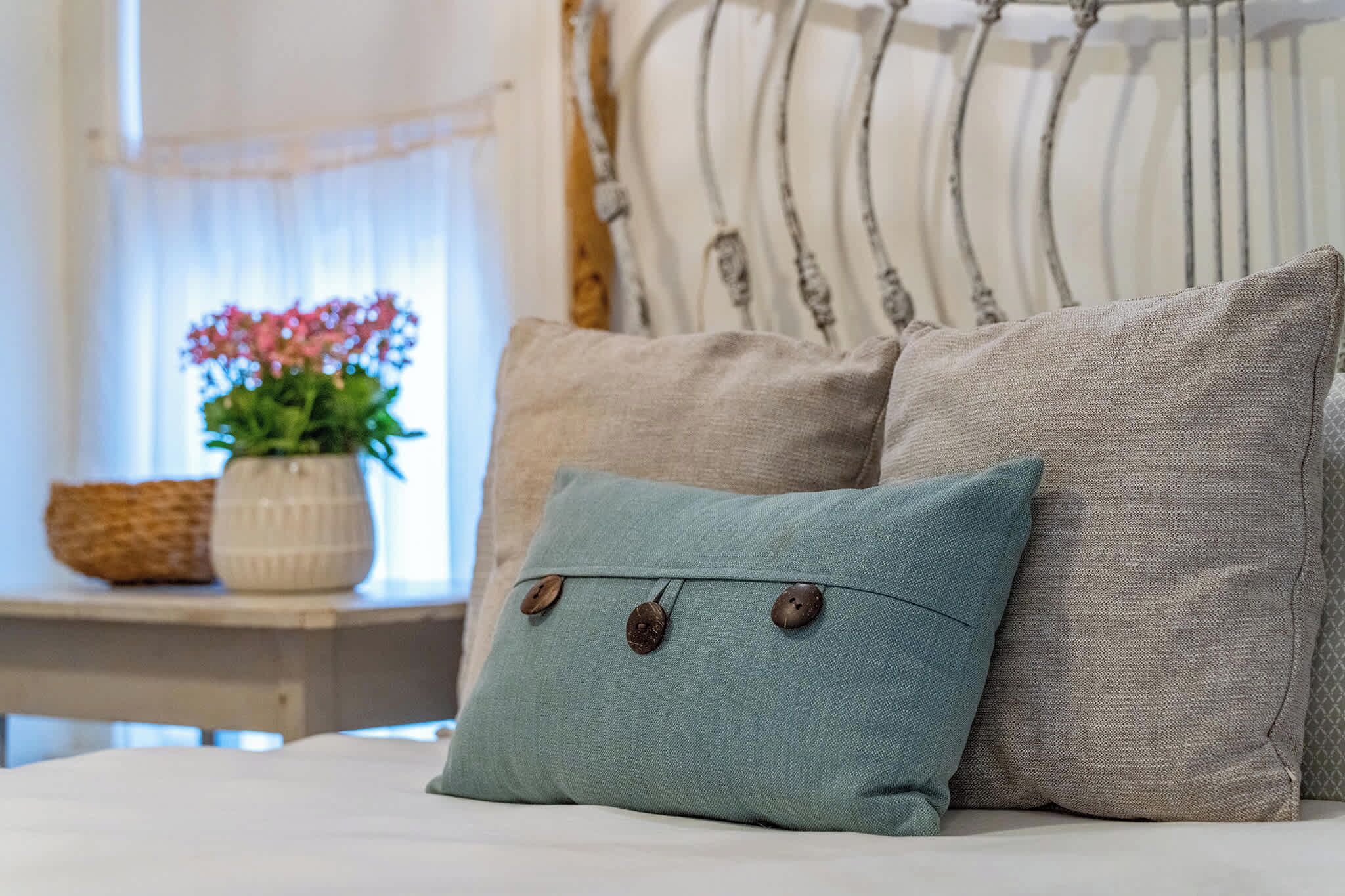 A bed with three pillows, a vase with flowers, and a window with curtains.