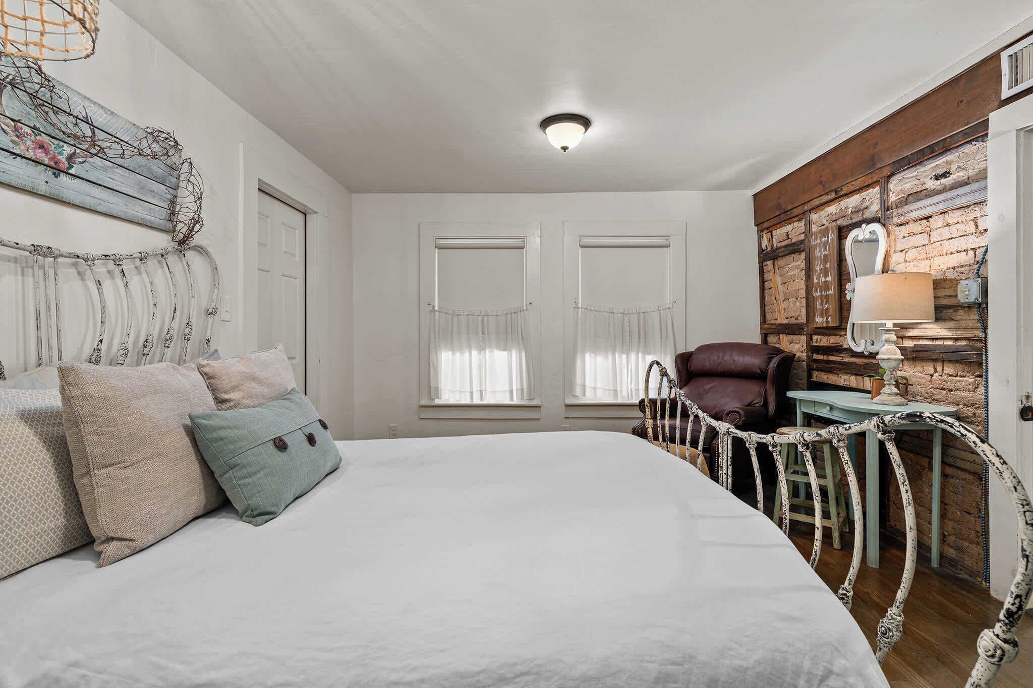 Une chambre avec un grand lit, deux fenêtres ornées de rideaux légers, une tête de lit en bois, un miroir décoratif et un mur au style rustique.