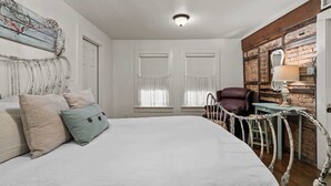 Un dormitorio con una cama grande, dos ventanas con cortinas translúcidas, un cabecero de madera, un espejo decorativo y una pared rústica.