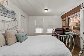 Ein Schlafzimmer mit einem großen Bett, zwei Fenstern mit durchscheinenden Vorhängen, einem Holz-Kopfteil, einem dekorativen Spiegel und einer rustikalen Wand.