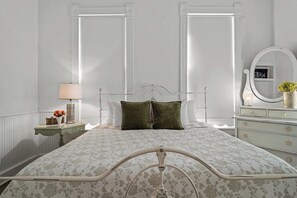 A bed with a metal frame, a nightstand with a lamp, a dresser with a mirror, and a wall with a window. - Ludwig Haus, Downtown, Pet Friendly, Fire pit! (New Braunfels)