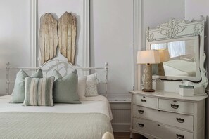 Une chambre à coucher avec un lit, des oreillers, une commode, un miroir et un luminaire fixé au mur.