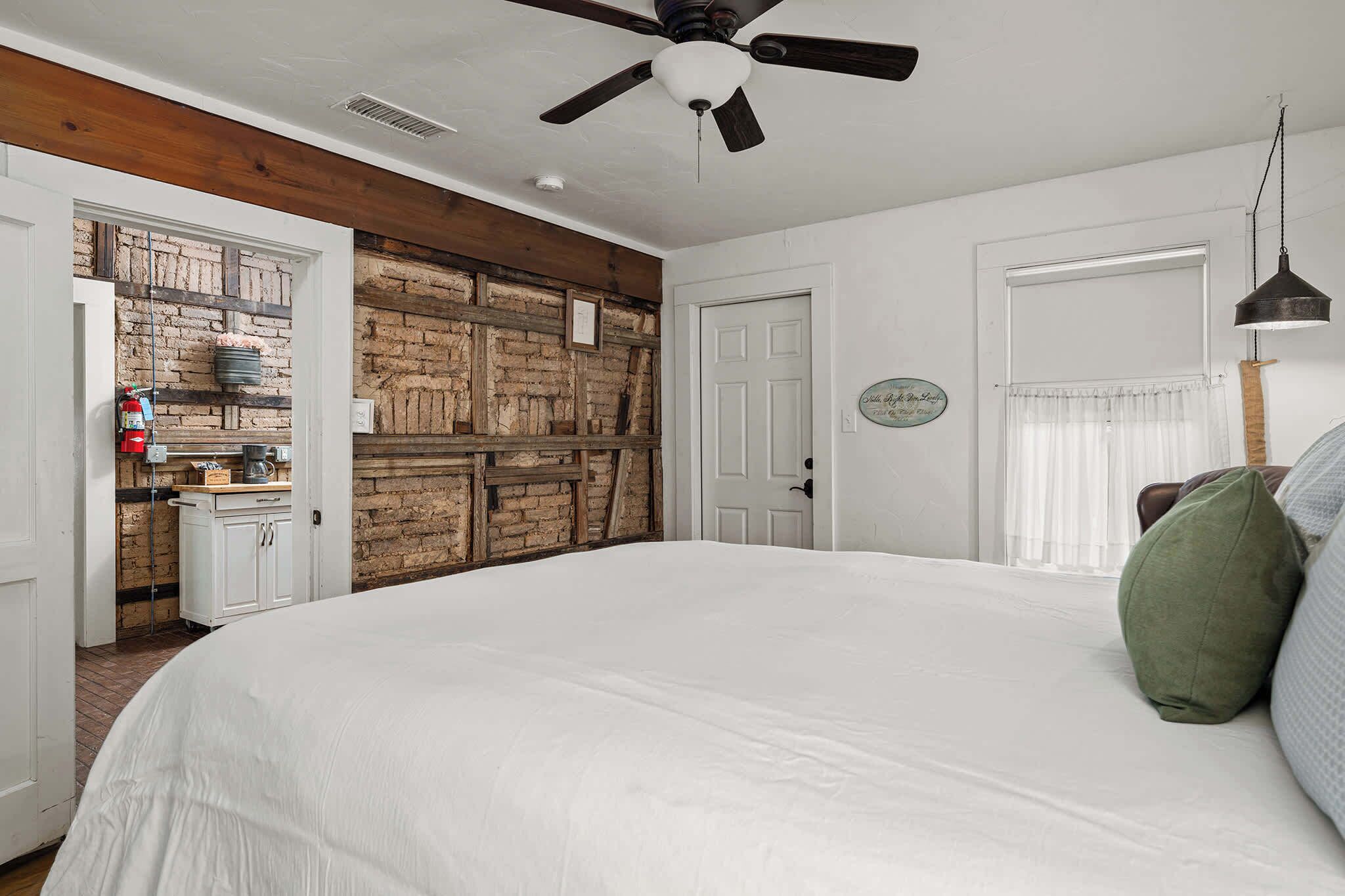 Quarto com uma cama grande, um ventilador de teto, uma parede de tijolos, uma porta e uma janela com cortinas.