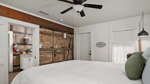 A bedroom with a large bed, a ceiling fan, a brick wall, a door, and a window with curtains. - Telegraph Station #1, Downtown, Pet friendly! (New Braunfels)