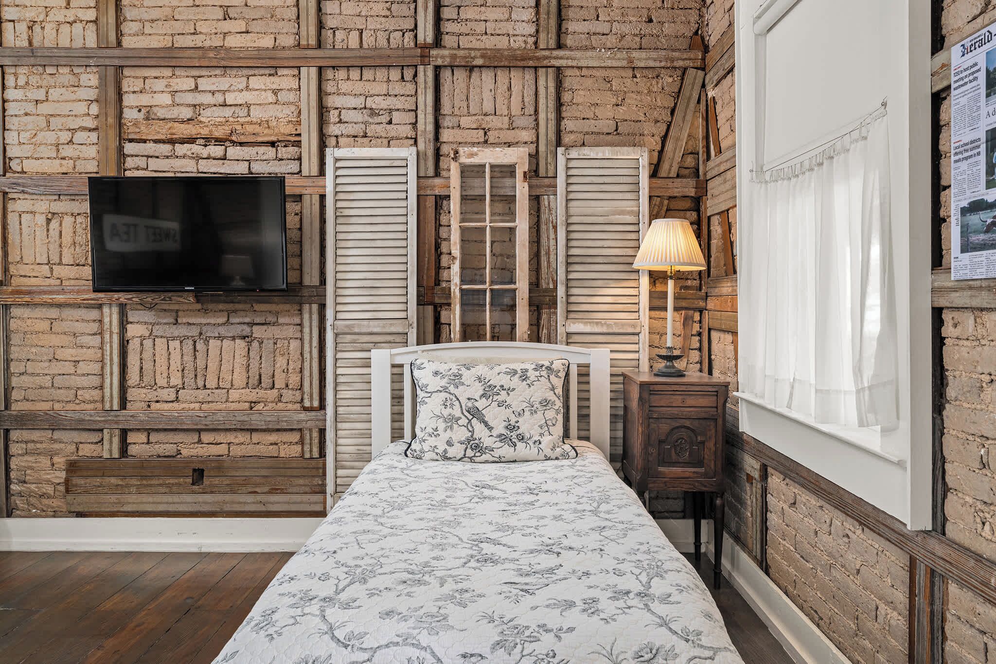 A bedroom with a brick wall, a bed with floral bedding, a television, and a window with curtains.