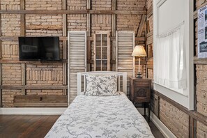 A bedroom with a brick wall, a bed with floral bedding, a television, and a window with curtains. - Telegraph Station #1, Downtown, Pet friendly! (New Braunfels)