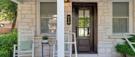 A stone house with a wooden door, two windows, a porch with white rocking chairs, and a small table with a plant.