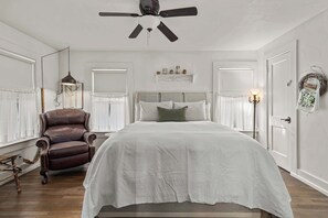 A bedroom with a bed, a leather armchair, a ceiling fan, and a nightstand with a lamp. - Telegraph Station #1, Downtown, Pet friendly! (New Braunfels)