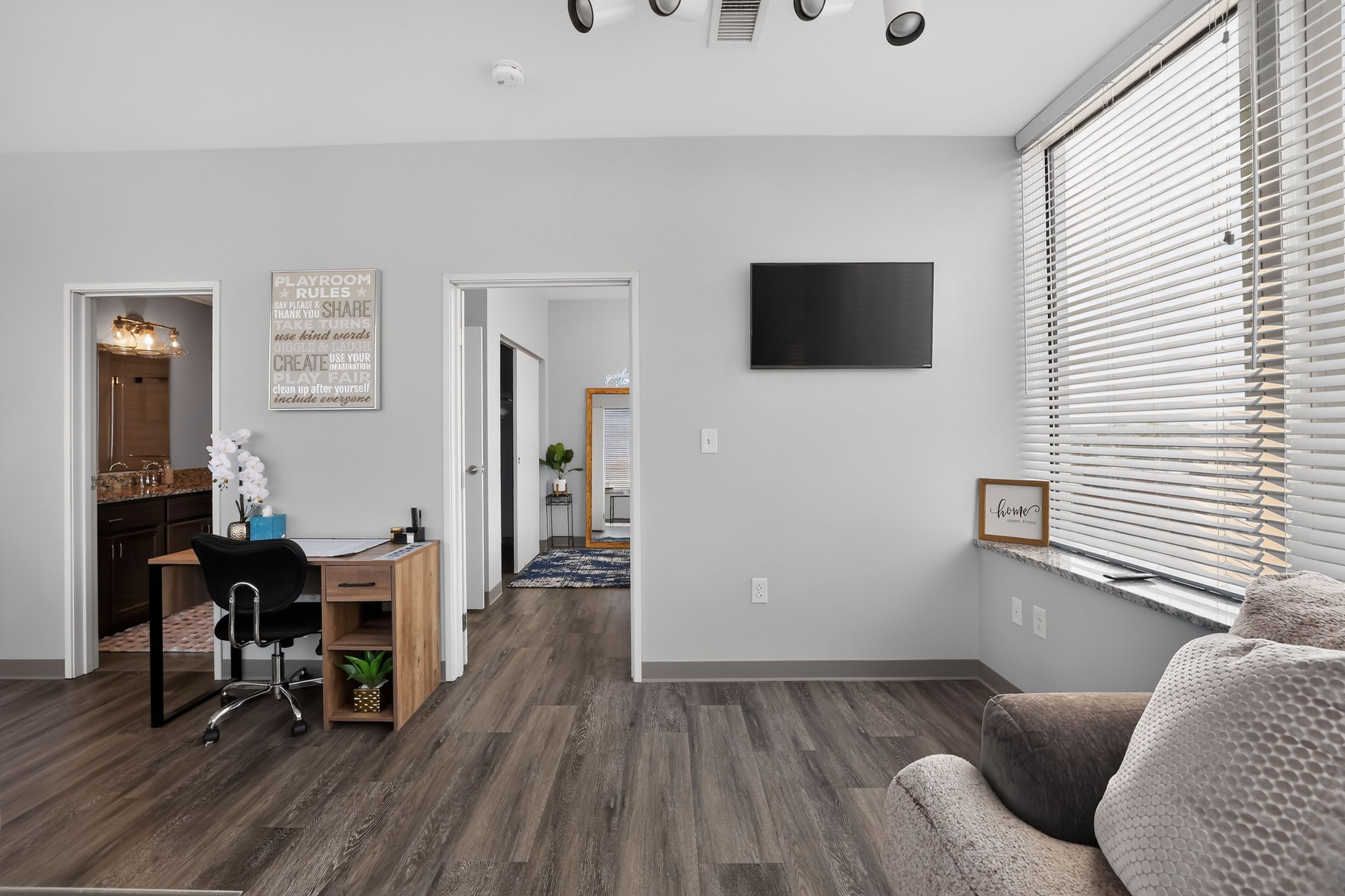 A modern living room with a wooden floor, a black TV mounted on the wall, and a large window with blinds.