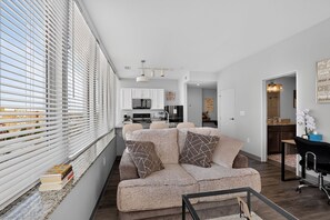 Ein modernes Wohnzimmer mit einer Couch, einem Glastisch und Blick auf die Küche durch große Fenster.