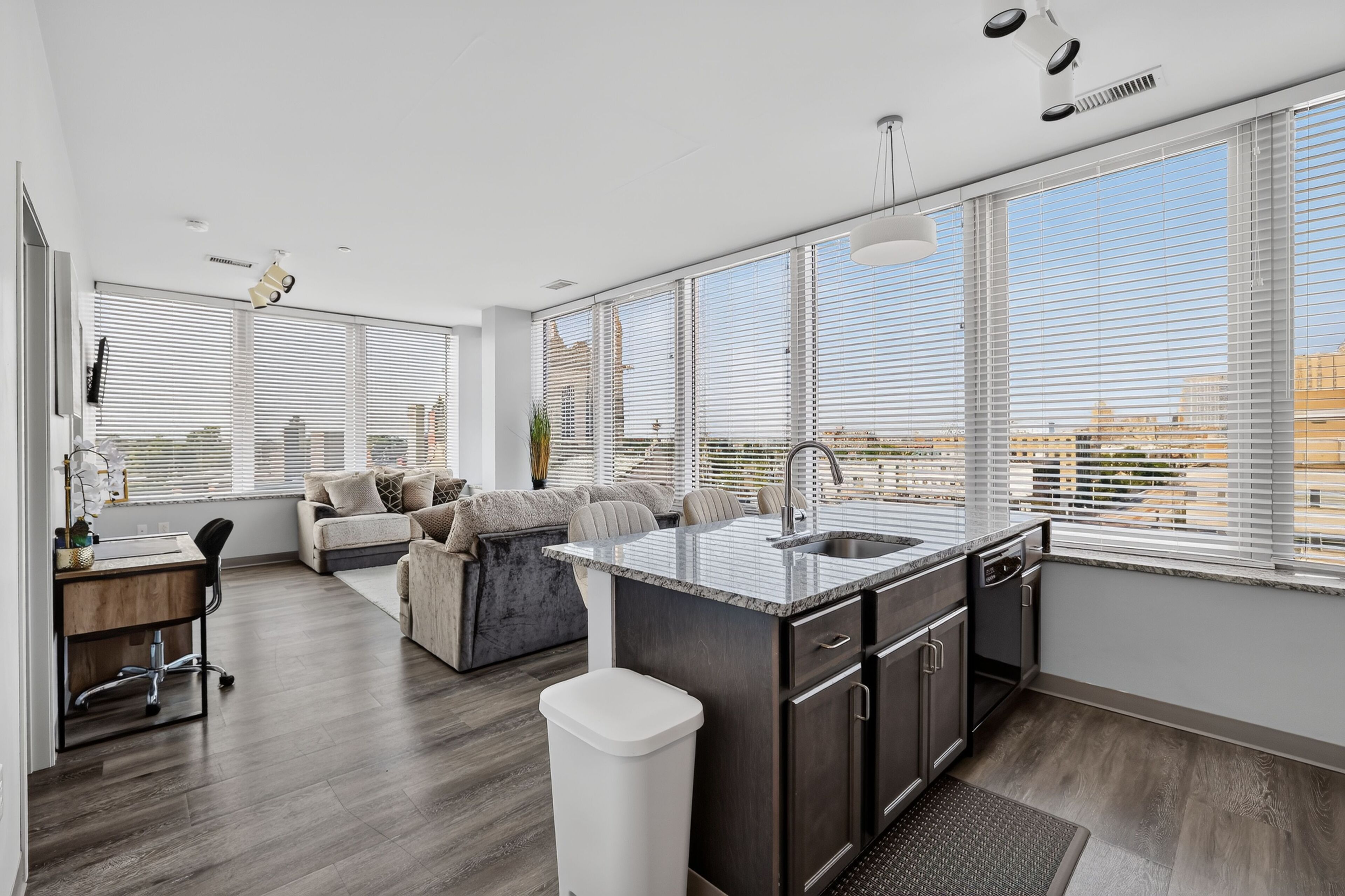 A modern kitchen with a large island, a dining area with a sofa, and large windows with blinds.