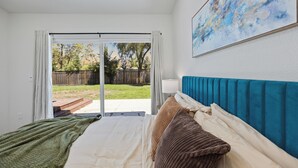 A bedroom with a bed, a nightstand, a lamp, a painting on the wall, and a view through a sliding glass door to the outside.