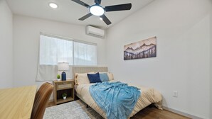 A bedroom with a bed, a desk, a chair, a ceiling fan, an air conditioner, and a window with curtains.