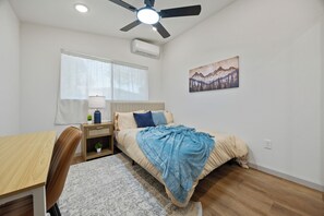 A bedroom with a bed, a desk, a chair, a ceiling fan, an air conditioner, and a window with curtains.