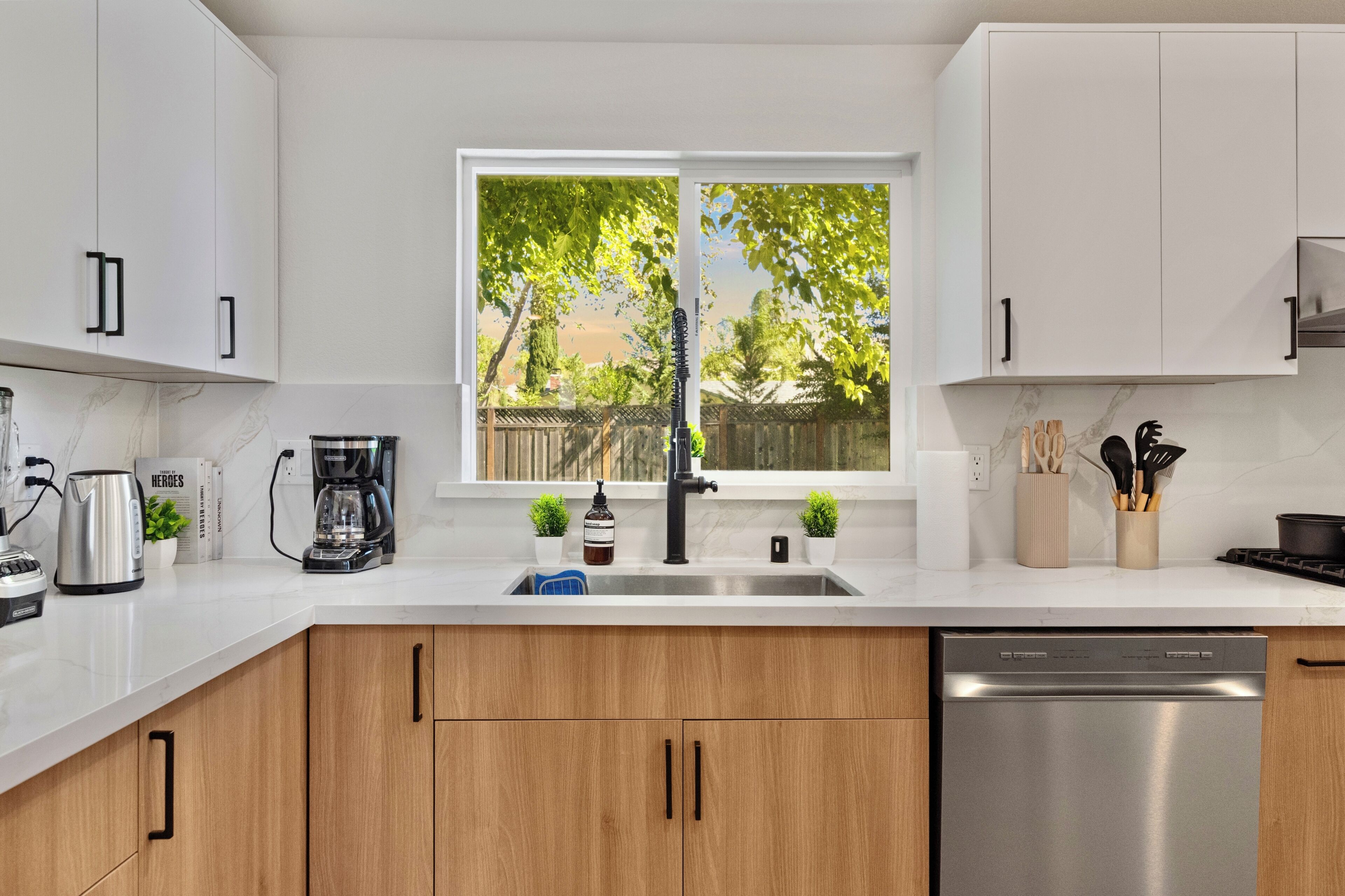 Cocina moderna con armarios blancos, lavavajillas de acero inoxidable y ventana con vistas a árboles.